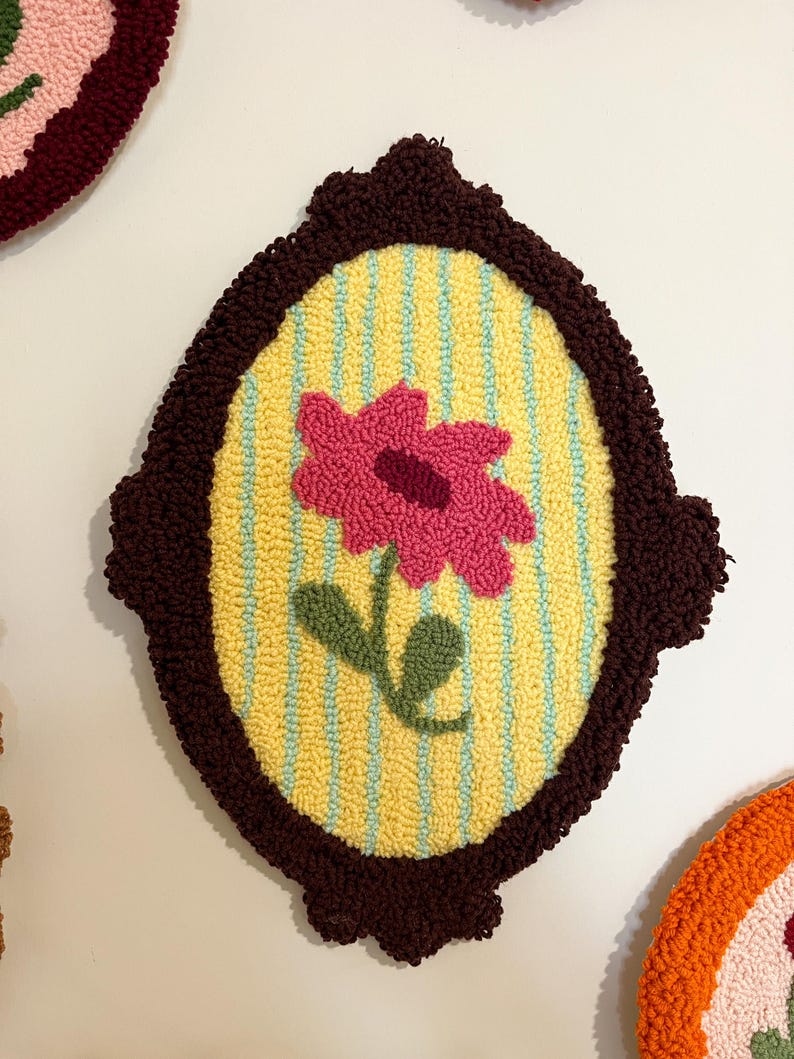 Pode incluir: Uma tape&ccedil;aria de parede oval feita &agrave; m&atilde;o com uma flor rosa e folhas verdes sobre um fundo amarelo com listras azuis. A flor &eacute; emoldurada por uma borda marrom escura texturizada. Outras pe&ccedil;as semelhantes s&atilde;o vis&iacute;veis.