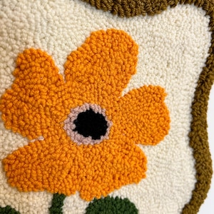 Pode incluir: Close-up de uma tape&ccedil;aria de parede artesanal e texturizada, com uma grande flor laranja com centro preto e detalhes rosa. A flor est&aacute; sobre um fundo creme, com caule e folhas verdes. A borda &eacute; marrom escuro.