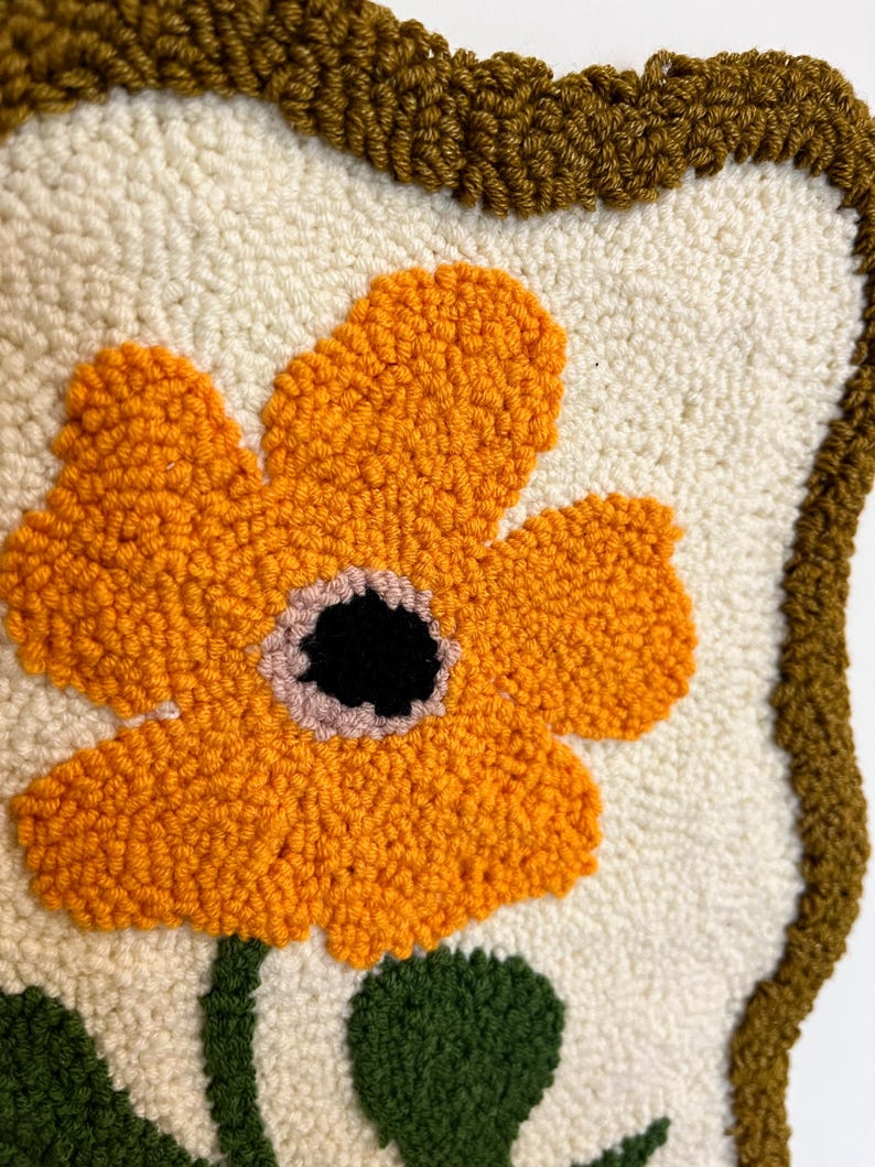 Pode incluir: Close-up de uma tape&ccedil;aria de parede artesanal e texturizada, com uma grande flor laranja com centro preto e detalhes rosa. A flor est&aacute; sobre um fundo creme, com caule e folhas verdes. A borda &eacute; marrom escuro.