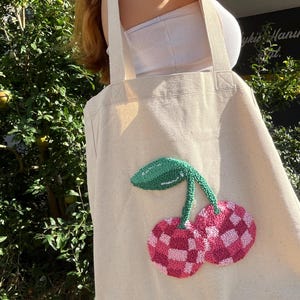 Op de afbeelding: Een witte canvas tote bag met een roze en groene kersenprint. De kers is geborduurd met een geruit patroon.
