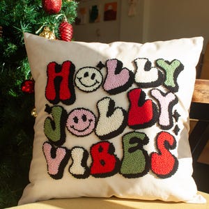May include: A white cushion with the words "HOLLY JOLLY VIBES" in colourful, puffy letters. The letters are outlined in black and have a smiley face in the middle of the "O" in "HOLLY" and "JOLLY".