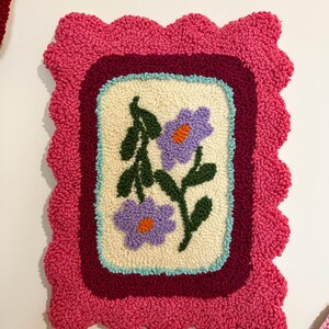 Pode incluir: Uma tape&ccedil;aria de parede retangular feita &agrave; m&atilde;o com um design floral. O painel central creme apresenta duas flores roxas com centros laranja e folhas verdes. O design &eacute; emoldurado por uma borda bord&ocirc;, uma borda azul clara e uma borda rosa ondulada.