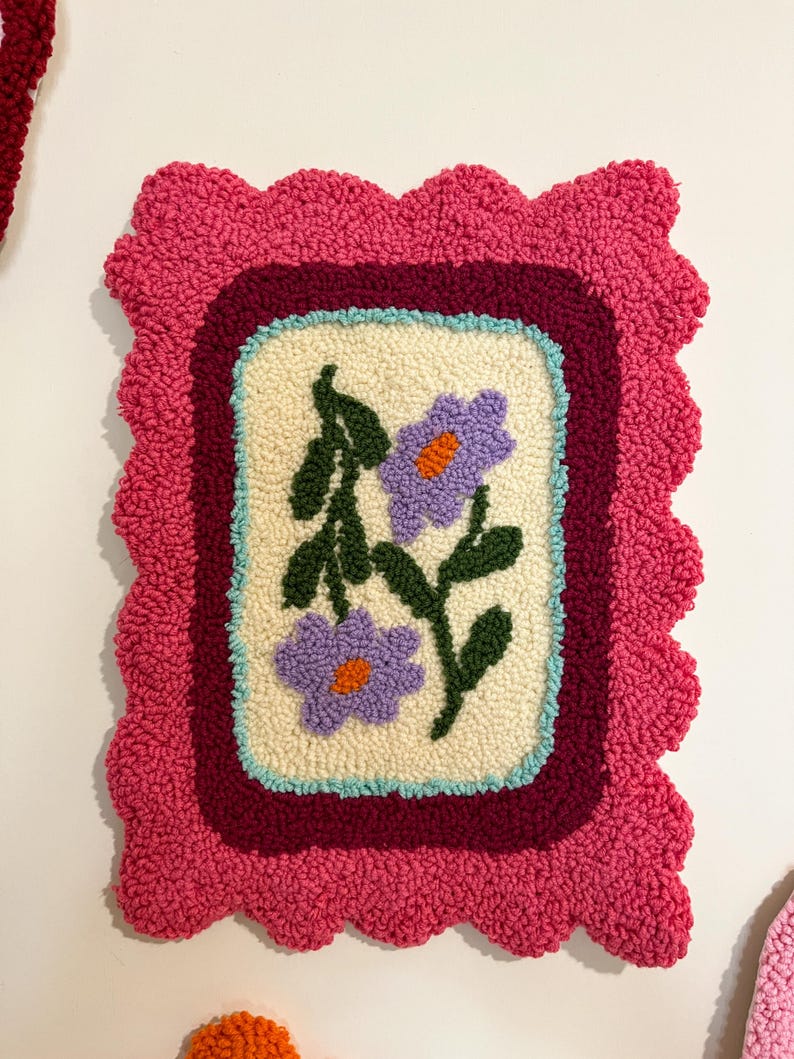 Pode incluir: Uma tape&ccedil;aria de parede retangular feita &agrave; m&atilde;o com um design floral. O painel central creme apresenta duas flores roxas com centros laranja e folhas verdes. O design &eacute; emoldurado por uma borda bord&ocirc;, uma borda azul clara e uma borda rosa ondulada.