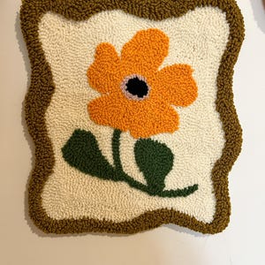 Pode incluir: Uma pe&ccedil;a de parede quadrada e texturizada com uma grande flor laranja com centro preto e folhas verdes. A flor est&aacute; sobre um fundo branco sujo, emoldurada por uma borda marrom. A pe&ccedil;a &eacute; provavelmente feita &agrave; m&atilde;o.