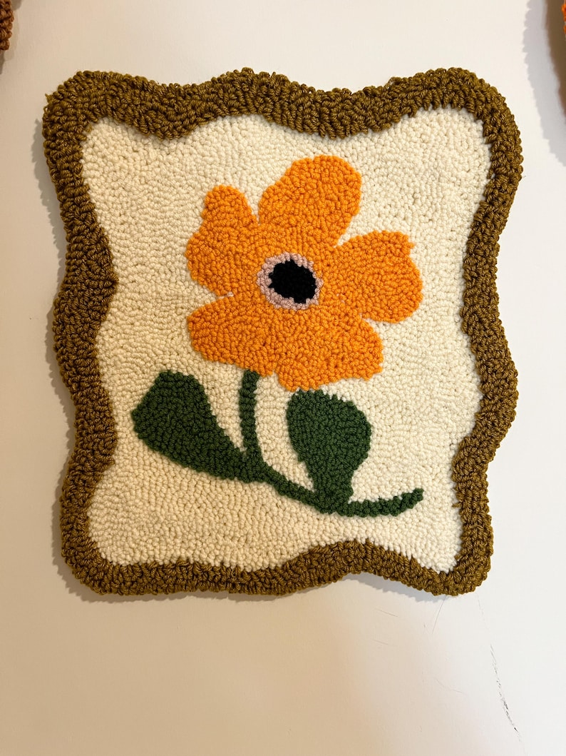 Pode incluir: Uma pe&ccedil;a de parede quadrada e texturizada com uma grande flor laranja com centro preto e folhas verdes. A flor est&aacute; sobre um fundo branco sujo, emoldurada por uma borda marrom. A pe&ccedil;a &eacute; provavelmente feita &agrave; m&atilde;o.