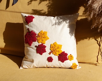 Peut inclure: Un coussin décoratif carré, blanc cassé, orné de feuilles d'automne brodées dans des tons de rouge et de jaune. Le coussin est posé sur un canapé jaune, créant une ambiance automnale chaleureuse. Les feuilles sont disposées en diagonale.