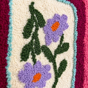 Pode incluir: Close-up de um t&ecirc;xtil texturizado e feito &agrave; m&atilde;o com um design floral. O ret&acirc;ngulo central creme apresenta duas flores roxas com centros laranja e folhas verdes. O ret&acirc;ngulo &eacute; emoldurado por uma borda azul clara, em um fundo rosa e bord&ocirc;.