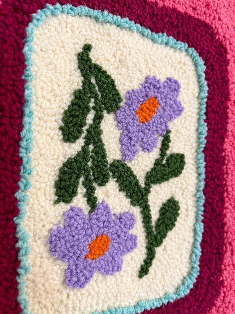 Pode incluir: Close-up de um t&ecirc;xtil texturizado e feito &agrave; m&atilde;o com um design floral. O ret&acirc;ngulo central creme apresenta duas flores roxas com centros laranja e folhas verdes. O ret&acirc;ngulo &eacute; emoldurado por uma borda azul clara, em um fundo rosa e bord&ocirc;.
