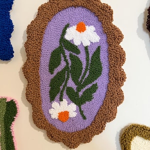Pode incluir: Decora&ccedil;&atilde;o de parede ovalada e texturizada com um design de margarida. Possui uma borda marrom ondulada, um interior lil&aacute;s e margaridas brancas com centros laranja e folhas verdes. A obra de arte &eacute; provavelmente feita &agrave; m&atilde;o.