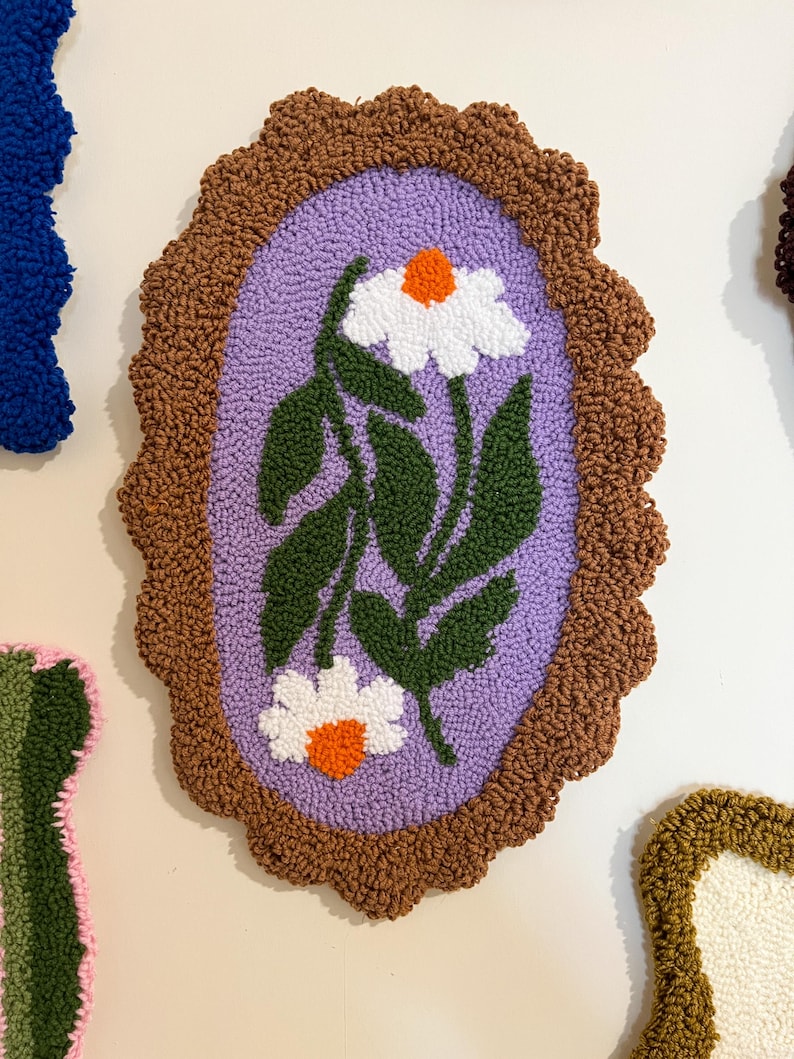 Pode incluir: Decora&ccedil;&atilde;o de parede ovalada e texturizada com um design de margarida. Possui uma borda marrom ondulada, um interior lil&aacute;s e margaridas brancas com centros laranja e folhas verdes. A obra de arte &eacute; provavelmente feita &agrave; m&atilde;o.
