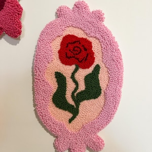 Pode incluir: Uma tape&ccedil;aria de parede oval rosa, tufada, com um desenho de rosa vermelha. A rosa tem folhas e caule verdes. O fundo &eacute; rosa claro e a borda &eacute; rosa mais escuro. A tape&ccedil;aria de parede est&aacute; em um fundo branco.
