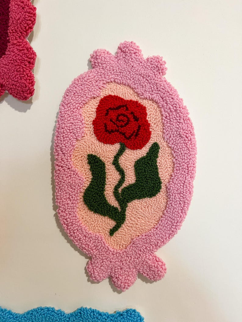 Pode incluir: Uma tape&ccedil;aria de parede oval rosa, tufada, com um desenho de rosa vermelha. A rosa tem folhas e caule verdes. O fundo &eacute; rosa claro e a borda &eacute; rosa mais escuro. A tape&ccedil;aria de parede est&aacute; em um fundo branco.