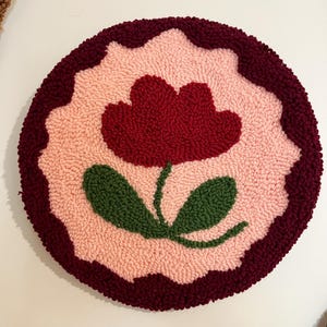 Pode incluir: Um tapete redondo e texturizado com um design floral. Apresenta uma borda vermelha escura, um fundo rosa claro e uma flor central vermelha escura com folhas verdes. O design &eacute; em estilo tufado, feito &agrave; m&atilde;o.