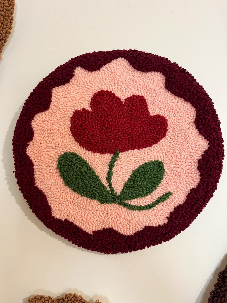 Pode incluir: Um tapete redondo e texturizado com um design floral. Apresenta uma borda vermelha escura, um fundo rosa claro e uma flor central vermelha escura com folhas verdes. O design &eacute; em estilo tufado, feito &agrave; m&atilde;o.