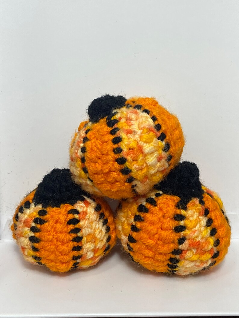 Pu&ograve; includere: Tre zucche all'uncinetto in arancione, giallo e nero. Le zucche sono impilate l'una sull'altra.