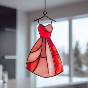Op de afbeelding: Een gebrandschilderd glazen jurk in rood- en roze tinten, hangend aan een metalen hanger. De jurk is samengesteld uit geometrische vormen en wordt opgehangen aan een ketting. De achtergrond is wazig, wat duidt op een binnenomgeving.