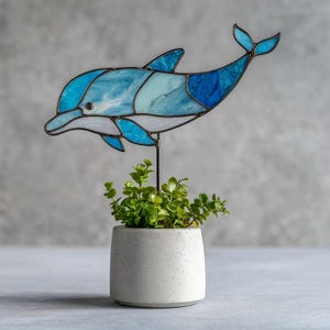 May include: A blue and white stained glass dolphin sculpture on a black metal rod, above a small, round, grey concrete planter with green plants. The dolphin is made of various shades of blue and white glass.