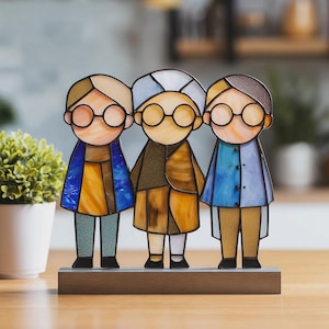 Könnte beinhalten: Ein Glasbild mit drei Figuren mit runden Brillen und farbenfroher Kleidung. Die Figuren stehen auf einem Holzsockel, mit Details in Blau, Gelb, Braun und Beige. Das Kunstwerk hat ein skurriles, handgefertigtes Aussehen.