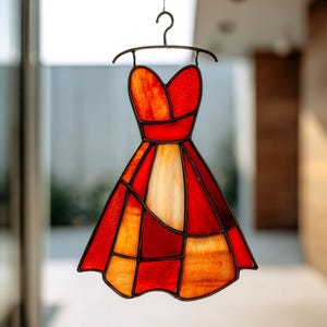 May include: A stained glass dress in shades of red, orange, and white, hanging from a metal hanger. The dress is composed of geometric shapes and has a strapless bodice and a flared skirt. The glass is translucent, allowing light to pass through.