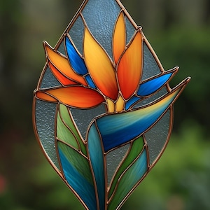 Könnte beinhalten: Buntglas-Kunstwerk in Diamantenform mit einer leuchtenden Paradiesvogelblume. Die Blume hat orange, gelbe und blaue Blütenblätter mit grünen und blauen Blättern. Das Stück ist mit einem kupferfarbenen Metall gerahmt.
