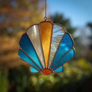 Puede incluir: Un atrapasoles de concha de cristal manchado con un marco de color cobre. La concha presenta secciones de vidrio blanco, dorado y azul. El atrapasoles está suspendido por una delicada cadena, perfecto para colgar.