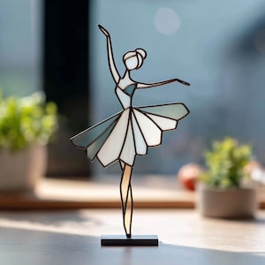 May include: A stained glass ballerina figurine in a graceful pose. The dancer's tutu is composed of geometric shapes in shades of white and blue. The figurine is mounted on a small black rectangular base, suitable for display on a shelf or table.