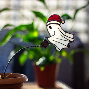 May include: A stained glass ghost wearing a red Santa hat, with a black wire stem, is attached to a potted cactus.