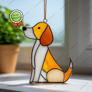 Op de afbeelding: Een gebrandschilderd glazen hondenornament in geel, oranje en wit. De hond zit, met een bruin ophangkoord. Het ornament heeft een "HOBY LICENCE" sticker. Het glas is donker omlijnd. Geschikt om op te hangen.