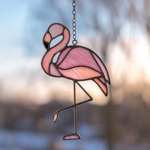 May include: A stained glass flamingo suncatcher in shades of pink and peach, with a black beak and metal frame. The flamingo is suspended by a chain, perfect for hanging in a window or on a wall. The design is a decorative piece.