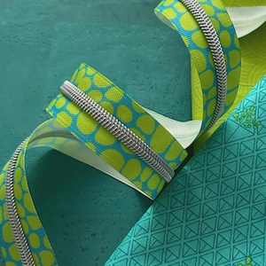 May include: Close-up of a zipper with a silver metal coil and a patterned fabric. The fabric features a repeating pattern of lime green and blue circles. A piece of teal fabric with a geometric design is also visible.