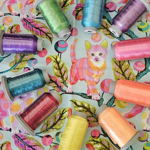 May include: A collection of colorful spools of thread arranged on a patterned fabric. The thread colors include green, blue, purple, pink, orange, red, and yellow. The fabric features a whimsical design with cats, leaves, and abstract shapes.