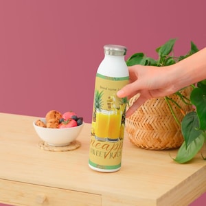 May include: White and silver insulated water bottle with a pineapple juice design. The label features a pineapple illustration and the words "pineapple sweet juice". The bottle is on a wooden table.