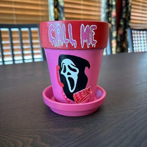 May include: A pink terracotta plant pot with a matching saucer, featuring a red rim with the text CALL ME in white dripping letters. The front displays a black and white ghost-like mask graphic with a red telephone handset and a red cord.