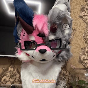 Może przedstawiać: Kolorowa głowa fursuit z wilczym designem. Głowa ma mieszankę szarego, niebieskiego, różowego i białego futra, z okularami w czarnych oprawkach i małą naszywką z misiem. Tekst "Fillndablanks" jest widoczny na dole.