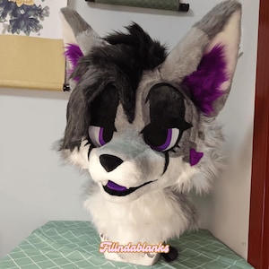 May include: A gray and white furry wolf head with purple ears and eyes. The head has a black nose and a purple mouth.