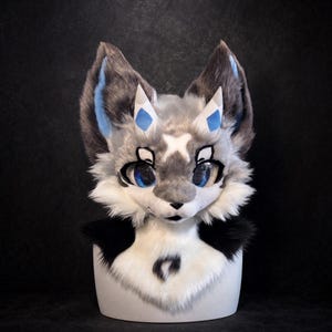 May include: A faux fur animal head mask featuring grey, white, and black fur. The mask has large blue eyes, pointed ears with blue inner accents, and small white horns with blue diamond markings. A white X-shaped mark sits on the forehead.