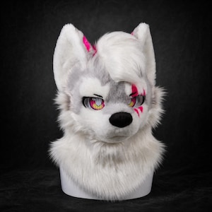 Può includere: Una maschera a testa di animale in peluche con pelliccia sintetica bianca e grigia, orecchie a punta con accenti rosa brillante e un naso nero. La maschera ha grandi occhi espressivi con iridi gialle e segni rosa intorno all'occhio.