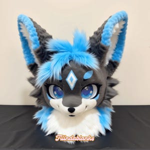 Puede incluir: Una cabeza de zorro de peluche con una cara gris y blanca, detalles en azul y grandes ojos azules. Las orejas y la frente presentan pelo azul brillante. El texto "Fillndablanks" está en la parte inferior.