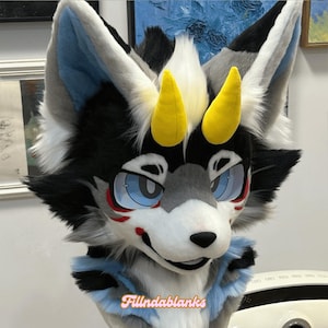 May include: A gray and white furry wolf head with yellow horns, blue eyes, and a red and white painted face. The wolf head is wearing a blue and white collar.