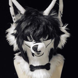 Peut inclure: Une tête de loup en fourrure noire et blanche avec un piercing et un nœud papillon. La tête de loup a des poils noirs sur le dessus et des poils blancs autour du visage et des oreilles. Les yeux sont gris et le nez est noir et blanc.