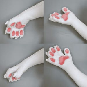 Pu&ograve; includere: Quattro immagini di zampe di peluche bianche con cuscinetti rosa. Le zampe sono in diverse posizioni, mostrando i dettagli del design. Il materiale in peluche appare morbido e soffice, con i cuscinetti rosa.