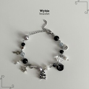May include: A Wybie bracelet with a silver chain, adorned with charms like a key, stars, a teddy bear, and beads in black, grey, and pearl. It fastens with a lobster clasp.