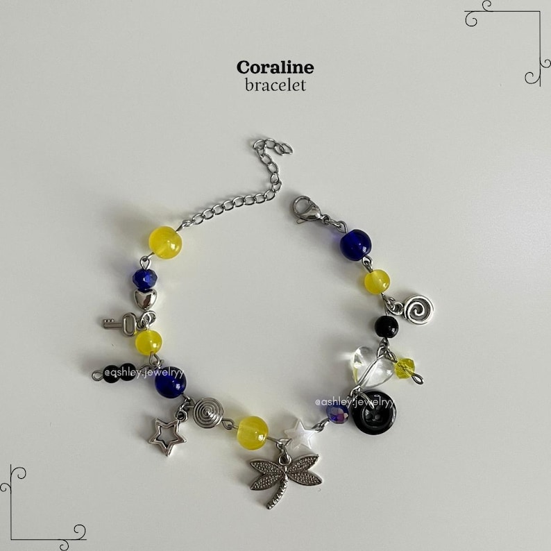 May include: A bracelet featuring yellow and blue beads, silver charms, and the text "Coraline bracelet." Charms include a key, star, dragonfly, and a spiral. The bracelet is displayed on a white surface.