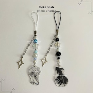 Puede incluir: Dos charms para teléfono con un diseño de pez betta blanco y negro. Cada charm tiene una cadena plateada con cuentas blancas y azules, una estrella plateada y un lazo de cuerda blanco o negro.