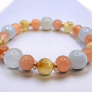 May include: A beaded bracelet featuring a variety of colorful gemstones. The bracelet includes white, orange, yellow, and peach-colored beads. The beads are strung together with gold-colored accents, creating a vibrant and eye-catching accessory.