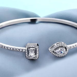 Puede incluir: Un brazalete de plata con una lágrima de cristal transparente y un cristal de forma cuadrada en los extremos. El brazalete está adornado con pequeños cristales transparentes.