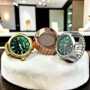 May include: Three ring watches with green watch faces and gold, rose gold, and silver bands. The watch faces display Roman numerals and the brand name "BOZHI". The rings are displayed on a white, textured surface.