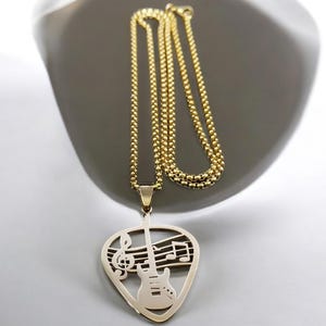 Puede incluir: Un collar de cadena dorada con un colgante de púa de guitarra. El colgante está recortado en forma de púa de guitarra con una guitarra y notas musicales en su interior.