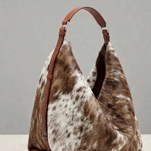 Könnte beinhalten: Eine Hobo-Schultertasche mit einem braun-weißen Kuhfellmuster und einer strukturierten, fellartigen Optik. Die Tasche verfügt über einen einzelnen verstellbaren braunen Lederriemen, der mit silberfarbenen Metallbeschlägen befestigt ist und eine lockere Form bildet.