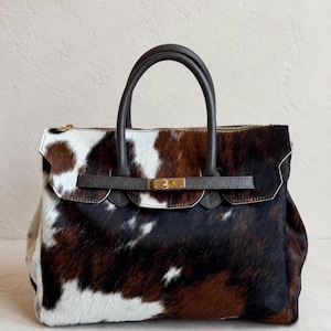 Könnte beinhalten: Eine Handtasche mit Kuhfellmuster in weißen, braunen und schwarzen Farbtönen. Die Tasche verfügt über zwei dunkelbraune Ledergriffe, einen horizontalen Lederriemen auf der Vorderseite und einen goldfarbenen Metallverschluss in der Mitte.